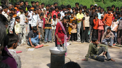 A large group of people gathered around a person with a metal bowl placed on a metal barrel. 