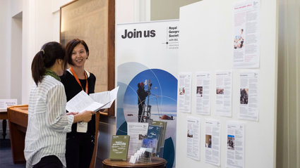 Two people chatting in front of a board of the Royal Geographical Society that says join us. 