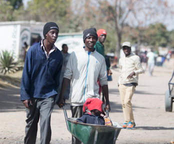 Two men walking along a dirt road pushing a child in a wheelbarrow