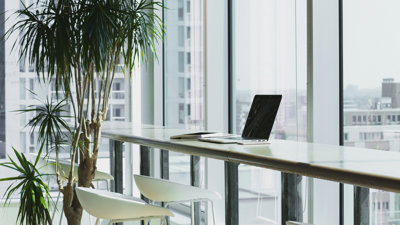 An open laptop on a table next to a wide set of tall glass windows in a modern office.