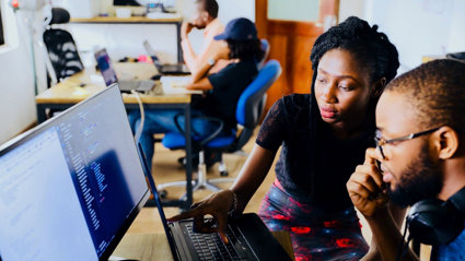 Two people sat at a desk in an office in front of a monitor and a laptop. One person is pointing to the laptop screen. There are other people working at a desk in the background.