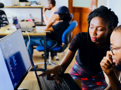 Two people sat at a desk in an office in front of a monitor and a laptop. One person is pointing to the laptop screen. There are other people working at a desk in the background.
