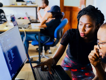 Two people sat at a desk in an office in front of a monitor and a laptop. One person is pointing to the laptop screen. There are other people working at a desk in the background.