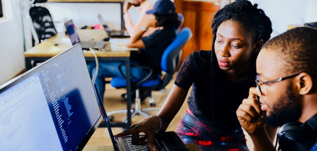Two people sat at a desk in an office in front of a monitor and a laptop. One person is pointing to the laptop screen. There are other people working at a desk in the background.