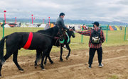 A rider on horseback, leading a second horse adorned with a red sash, receives a letter from a person standing on the ground.