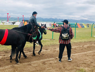 A rider on horseback, leading a second horse adorned with a red sash, receives a letter from a person standing on the ground.