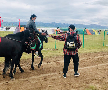 A rider on horseback, leading a second horse adorned with a red sash, receives a letter from a person standing on the ground.