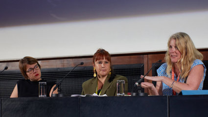 Three speakers on a conference panel sat behind a desk on a stage.
