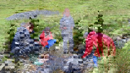 Three teachers sitting on the banks of a small, fast flowing stream and two standing in the water.