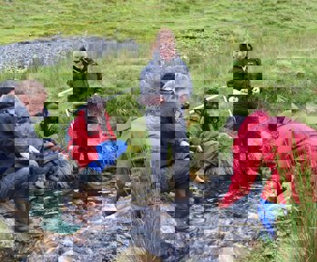 Three teachers sitting on the banks of a small, fast flowing stream and two standing in the water.