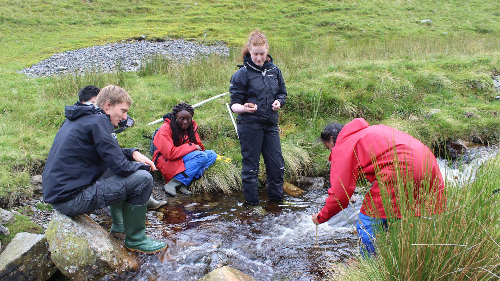 Three teachers sitting on the banks of a small, fast flowing stream and two standing in the water.