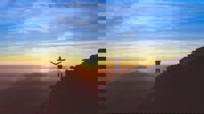 Person standing on a cliff with arms outstretched, in front of a sunset sky
