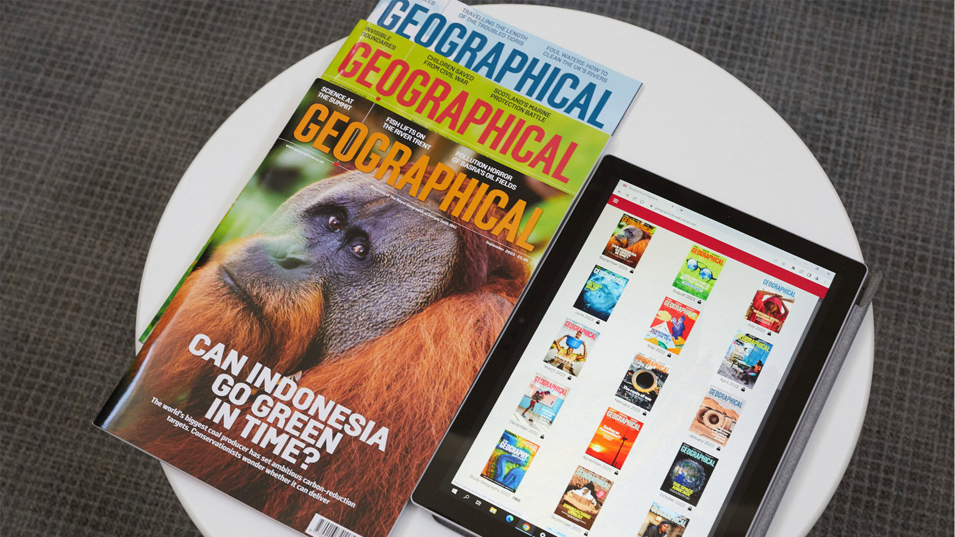A pile of three print copies of Geographical magazine next to a tablet that displays covers of magazines in the Geographical magazine app.