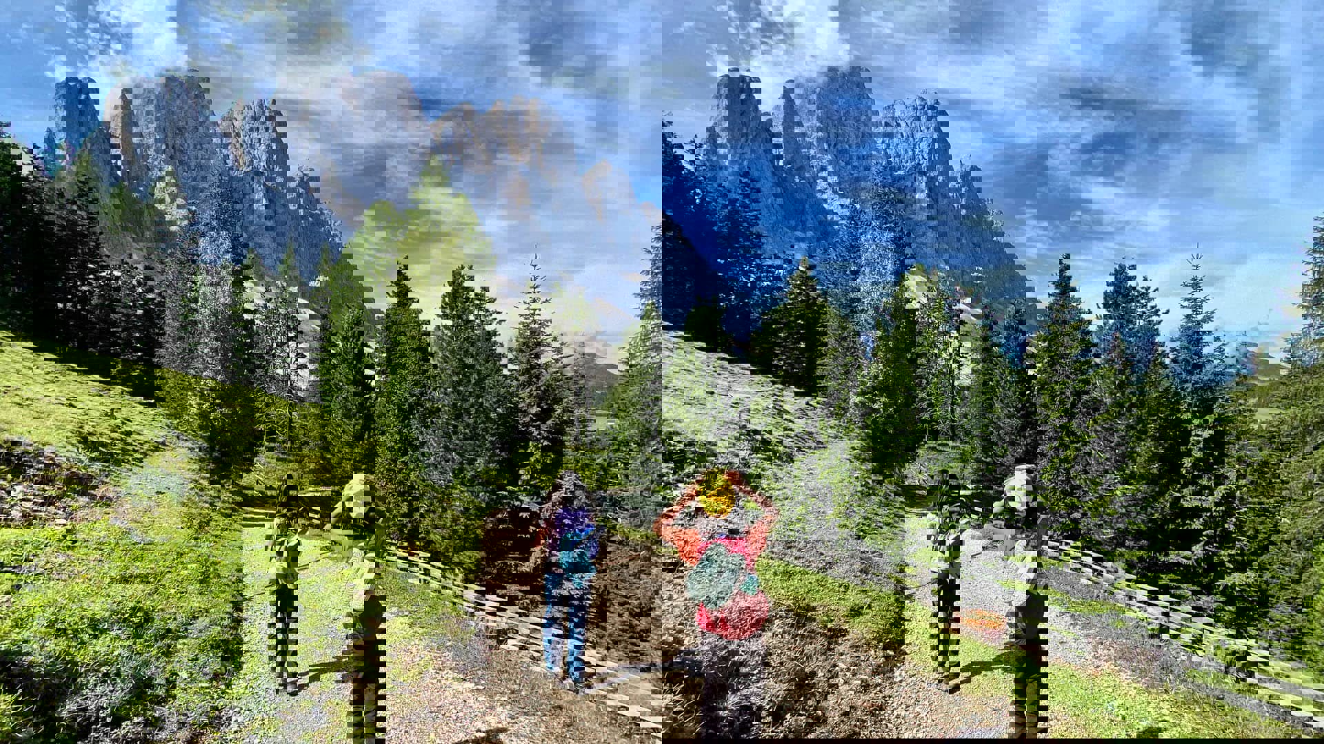 Two people hiking a mountain trail through the Italian Dolomites with tall alpine peaks in the background.
