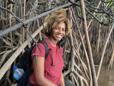 Woman in red t-shirt and wearing a backpack sitting at the edge of mangroves.