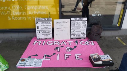 Pink cloth on sidewalk with 'Migration is Life' message and flyers about immigration raids and important info.