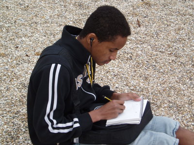 A boy sitting on a pebble beach, making notes in a notebook