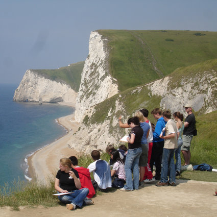A group of students are sitting near the edge of a cliff, with a view down onto a sandy beach with white chalk cliffs below them.