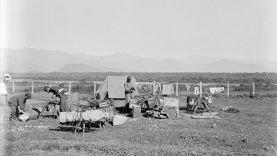 Four people camping in a vast field, in Fort Hertz, North Burma. Mountains can be seen far in the background. This visual is not in colour. 