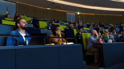 Audience members attentively listening in a modern lecture hall with tiered seating and green chairs.
