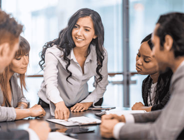 Four people sat around table in corporate wear, with a fifth person standing and pointing at a piece of paper on the table.