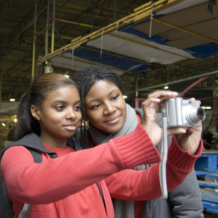 Two female students are in a market area, one is holding a digital camera and taking a picture of the scene. The other is looking at the picture being taken through the camera screen.