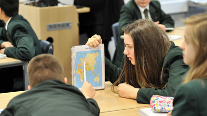 Children in a classroom. One holds a notebook with a world map, while another points at the map.
