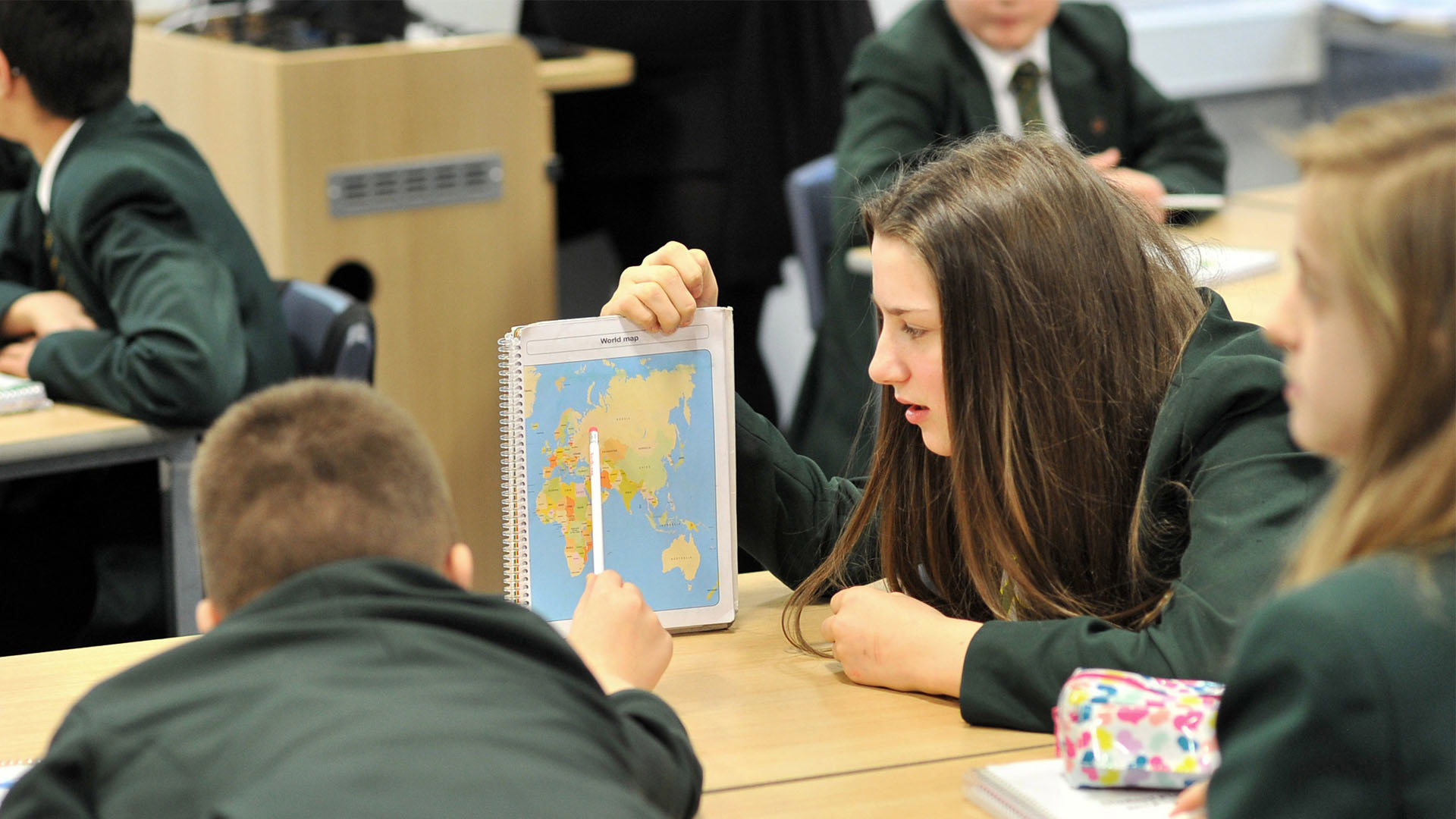 Children in a classroom. One holds a notebook with a world map, while another points at the map.