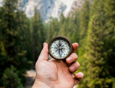 Hand holding a compass in front of mountain and forest background.