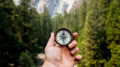 Hand holding a compass in front of mountain and forest background.