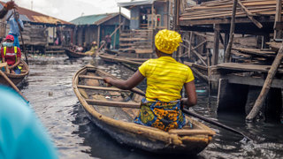 Person in a boat rowing through wooded buildings on stilts in the water.