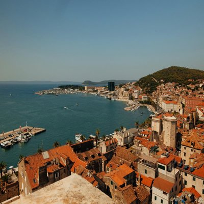 A view over a Mediterranean town by the sea. The roofs are orange tile and several jettys for boats can be seen
