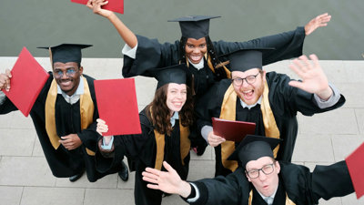 Group of five people in graduation gowns celebrating and raising their certificates.