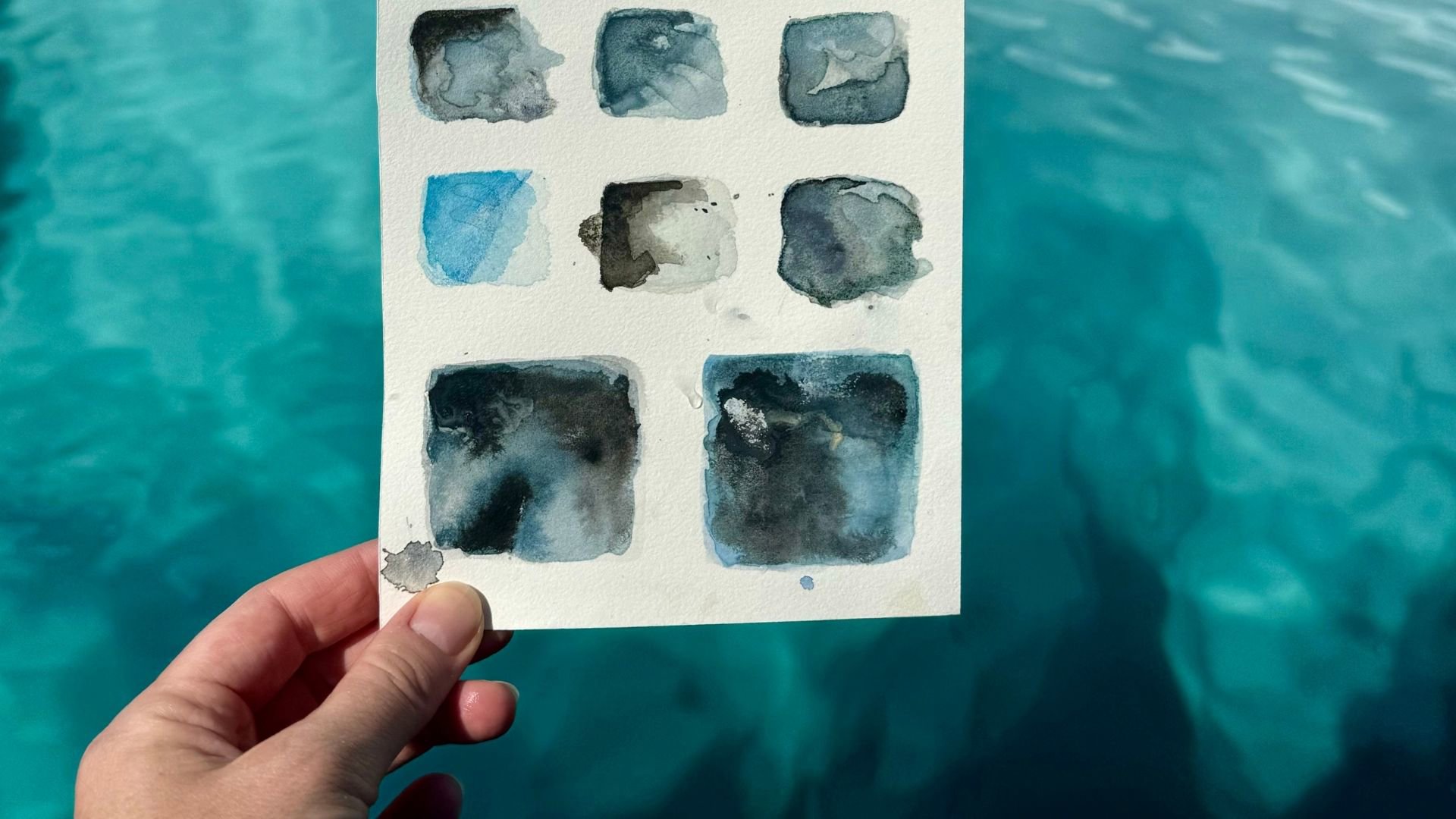 Hand holding a watercolour painting of water with the sea behind. 