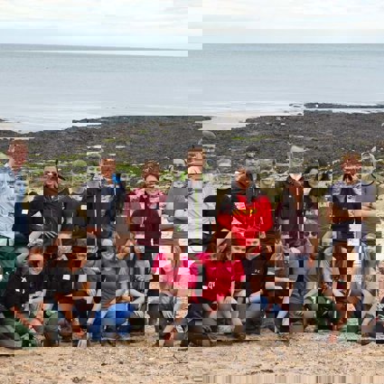 A group of older students lined up on a beach. In the background you can see a rocky beach with seaweed, and then the sea.
