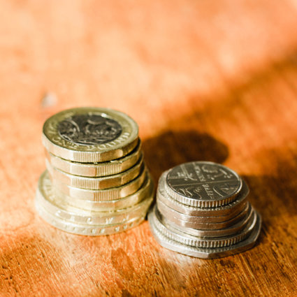 Two piles of english coins, pound coins and two pound coins to the left hand side and fifty pence, twenty pence and five pence to the right hand side, They are placed on a wooden table.