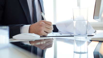 A person in a navy blue suit, white shirt and navy blue tie sits at a desk whilst using a pen to write on a spice of paper. A glass of water and white computer mouse sits next to him on the table.
