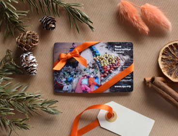 A membership card wrapped up in orange ribbon on brown wrapping paper with festive foliage surrounding it, including acorns, holly and cinnamon sticks.