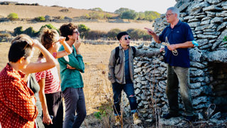 A person with a rope standing in front of a stone structure and speaking to a group of people in a dry, rural landscape.