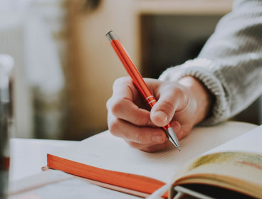 Person holding a pen and writing in an open notebook on a table.