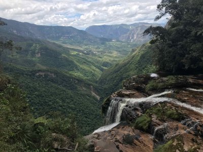 An evelated view of rocks hiding a water fall cascading into the large expanse of a rainforest.