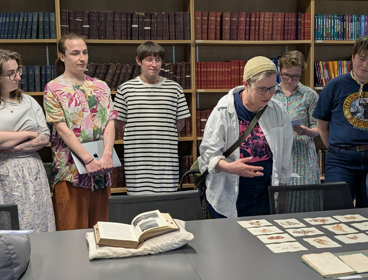 Six people standing up around a table and observing some Collections items.