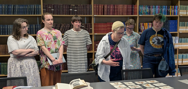 Six people standing up around a table and observing some Collections items.