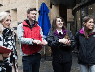 Four people outside a building, holding clipboards while smiling at something off-camera.