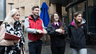 Four people outside a building, holding clipboards while smiling at something off-camera.