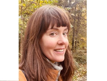 Person with long brown hair standing in a forest with autumn foliage in the background.