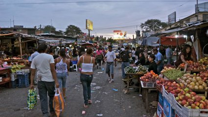People walking through an outdoor market with fresh fruit and vegetables displayed in crates on market stalls. 