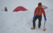 Expeditioner in winter gear caught in a snowstorm standing next to an improvised camp in a landscape covered in snow.