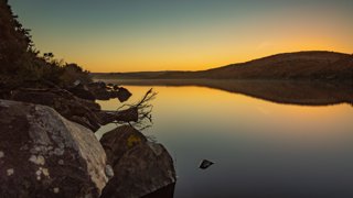 A wide and calm lake at sunset with rocks and fallen trees in the front.