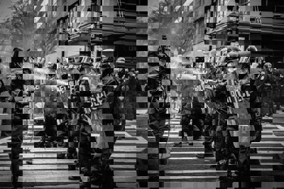 A wall of police officers with hemelts and bullet proof gear hold sheilds on front of themelves  a one of the officers uses sprays water at civilians which cant be seen form the angle.